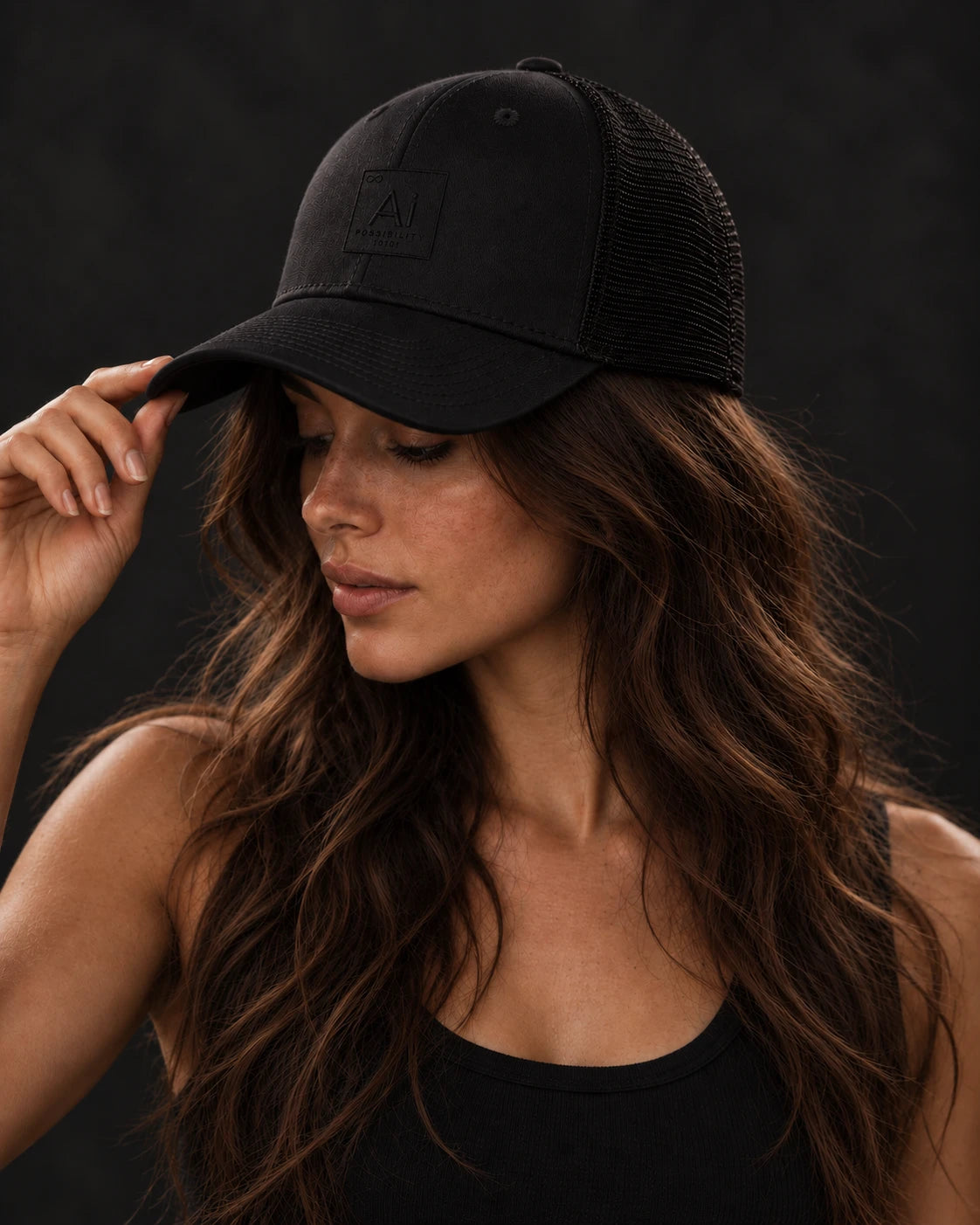 Woman wearing the Possibility Trucker Hat in black, hand on the brim, embroidered Ai element tile on the front, dark studio